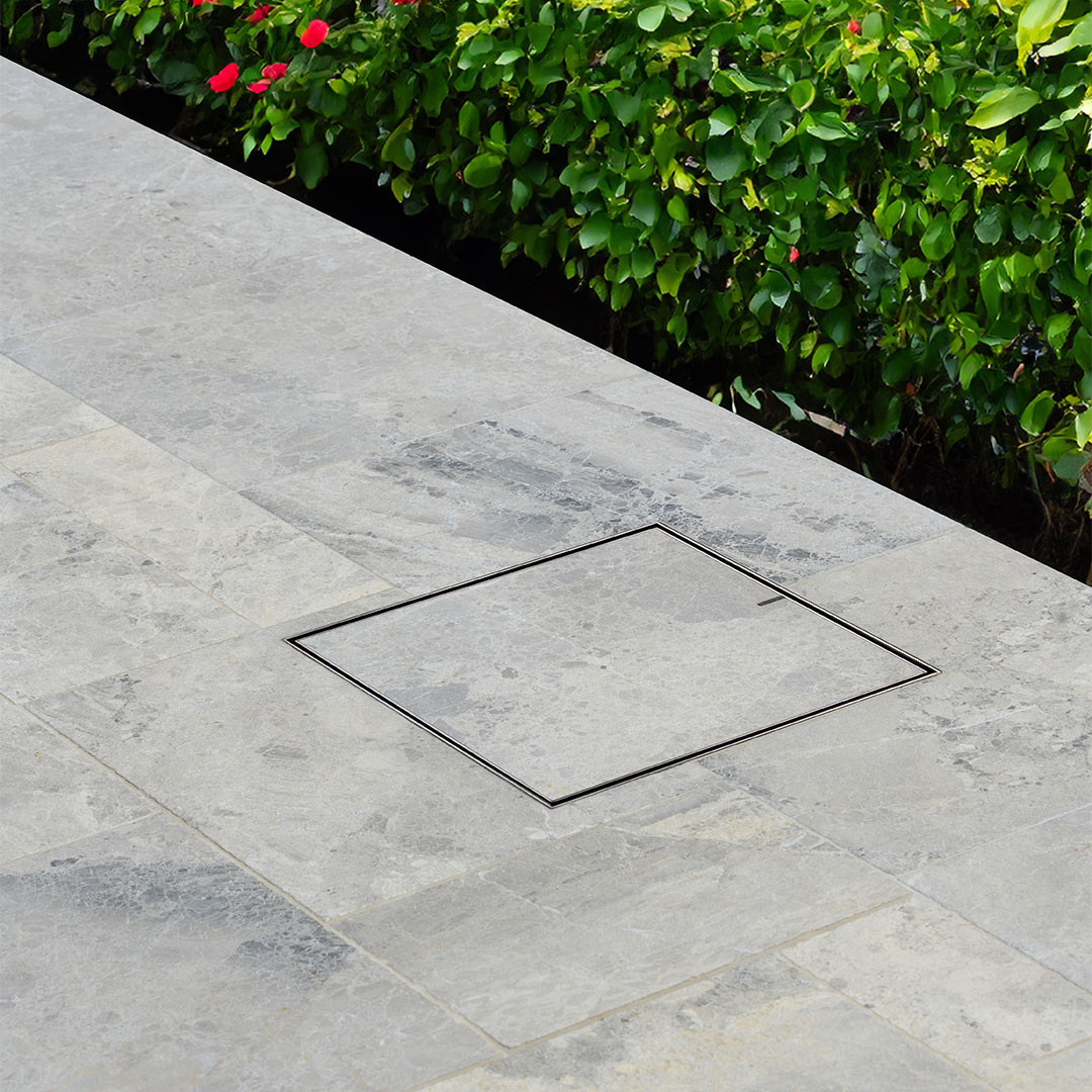 Square HIDE Drain Cover in a stone patio with greenery in the background.